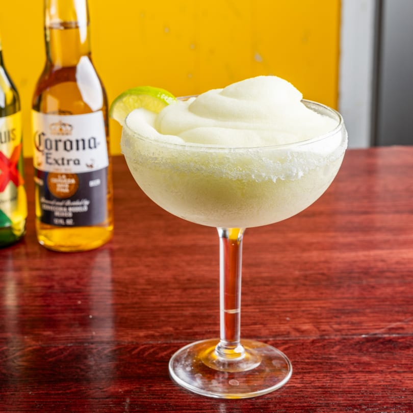 A frozen margarita with a lime wedge and salted rim, with two Corona bottles in the background.